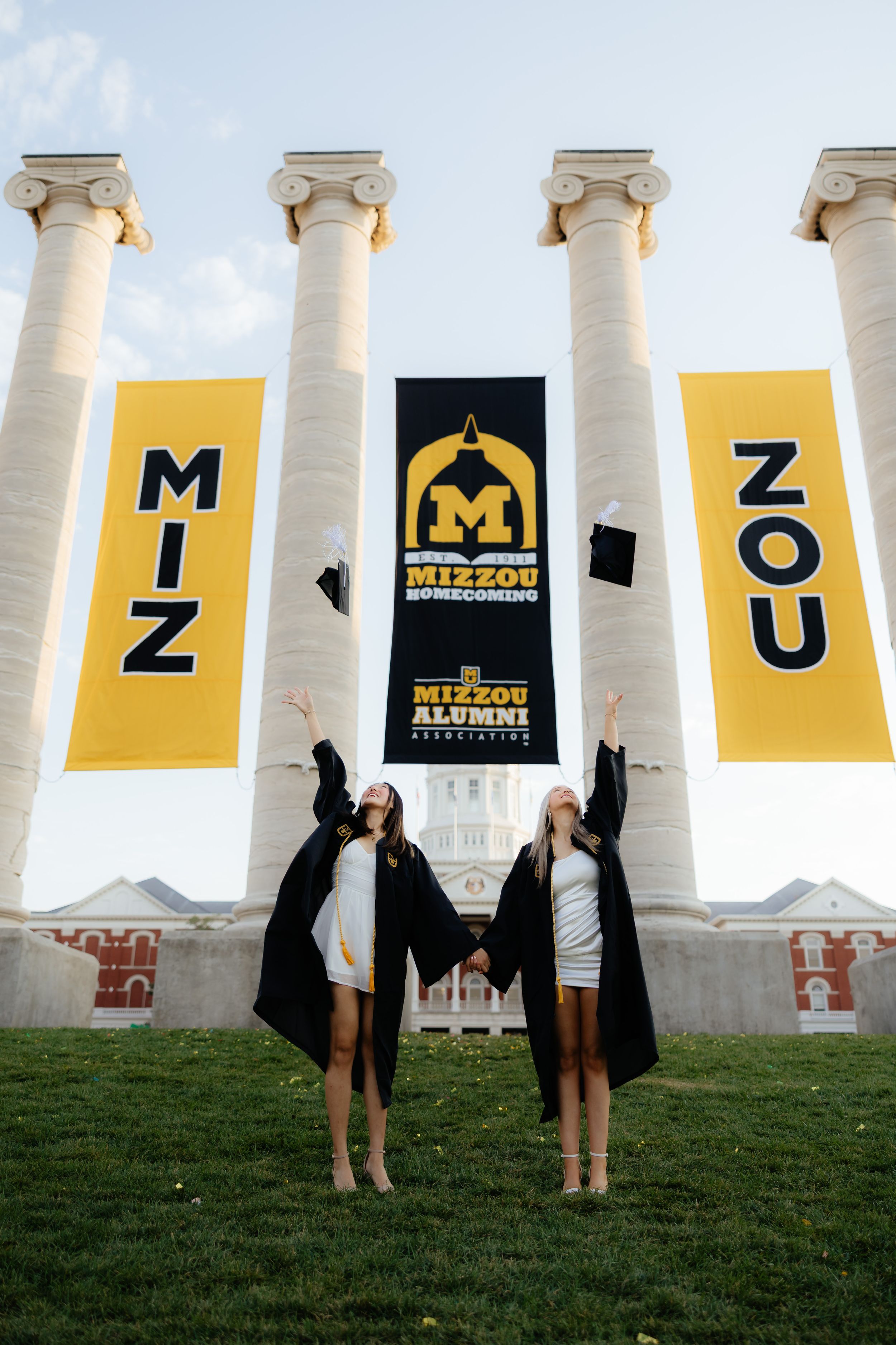 Two graduates jumping in celebration in front of Mizzou columns with yellow banners.