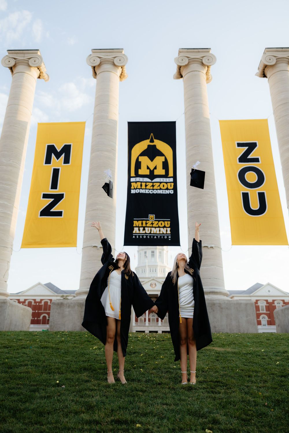 Two graduates jumping in celebration in front of Mizzou columns with yellow banners.