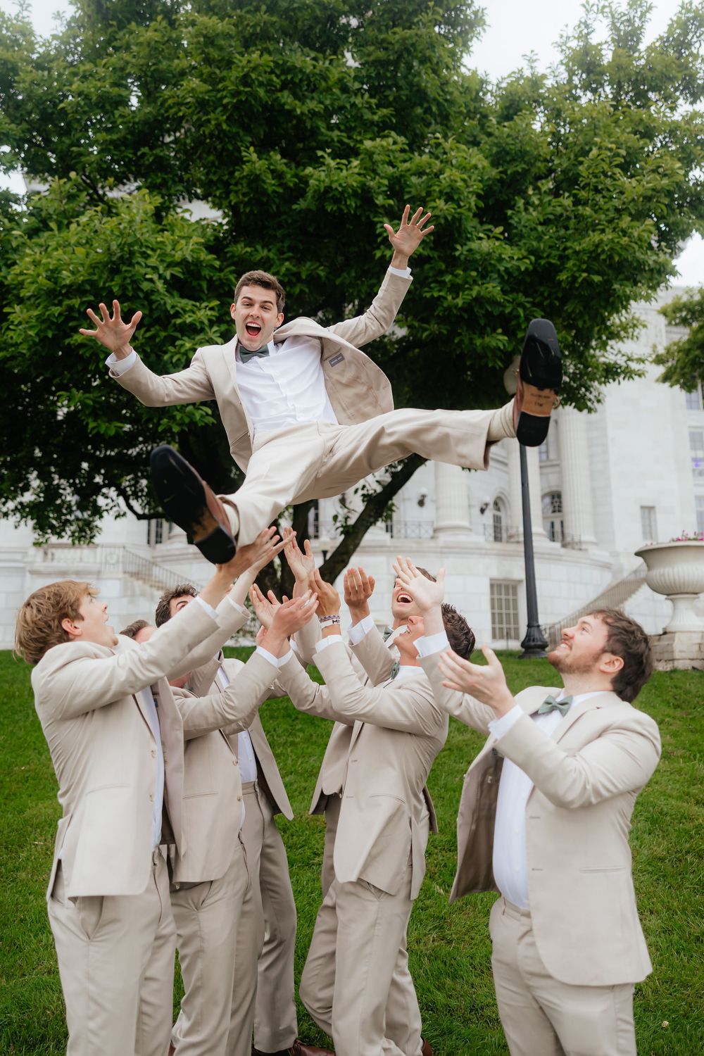 Groomsmen in light suits toss groom in air during playful wedding photo.