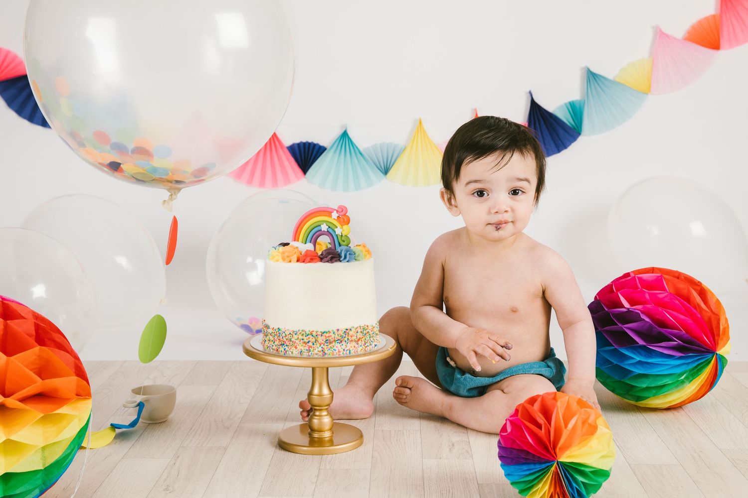 Colorful first birthday cake smash setup with rainbow decorations, honeycomb balls and festive bunting on light wood floor.