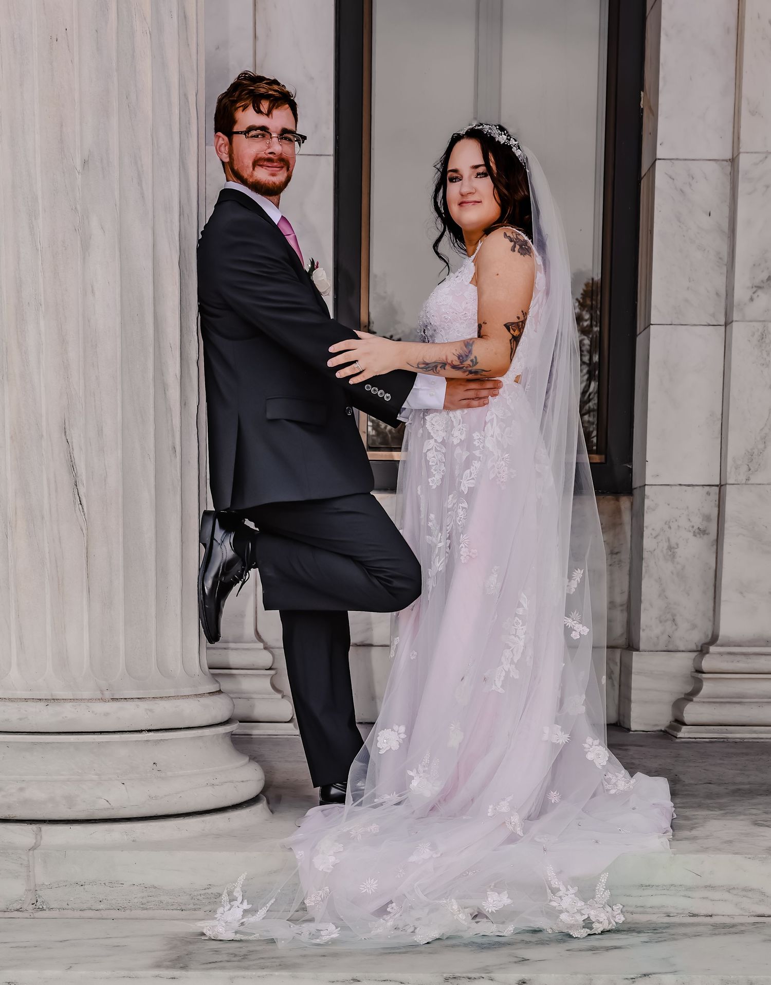 Romantic wedding couple poses playfully between marble columns in an elegant venue.