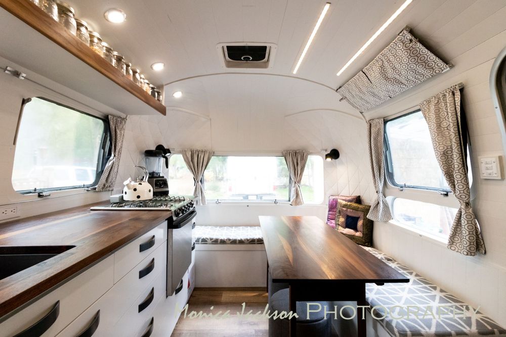 Bright modern Airstream trailer interior with wood counters, white cabinets, and cozy seating area.