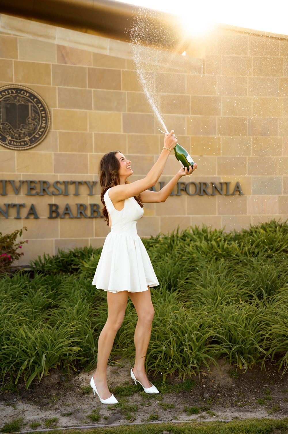 UCSB Grad portraits at Henley Gate during sunset of sorority senior in white dress spraying champagne