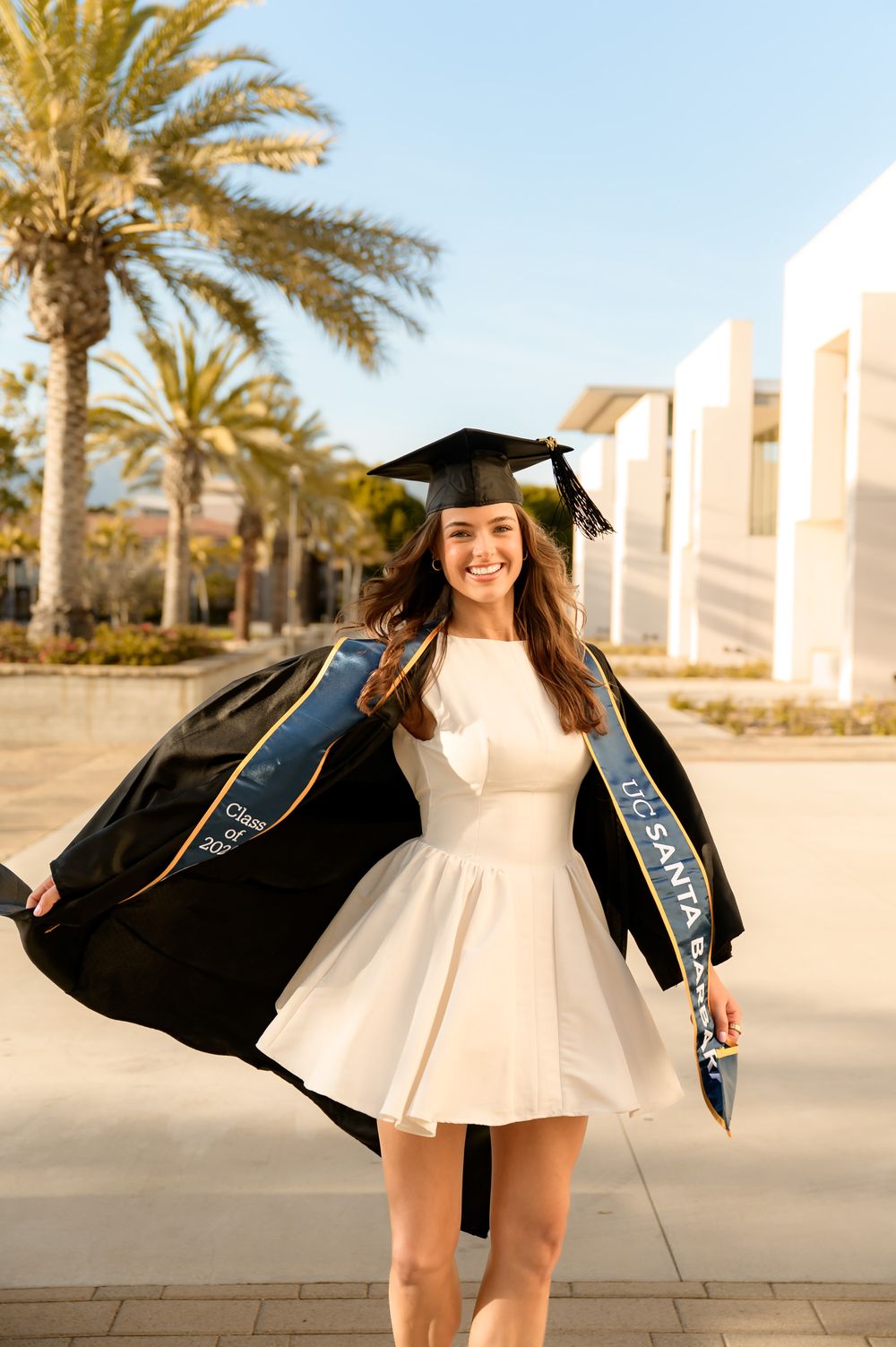 UCSB Grad portraits at library of sorority senior in white dress spinning and smiling in her cap and gown
