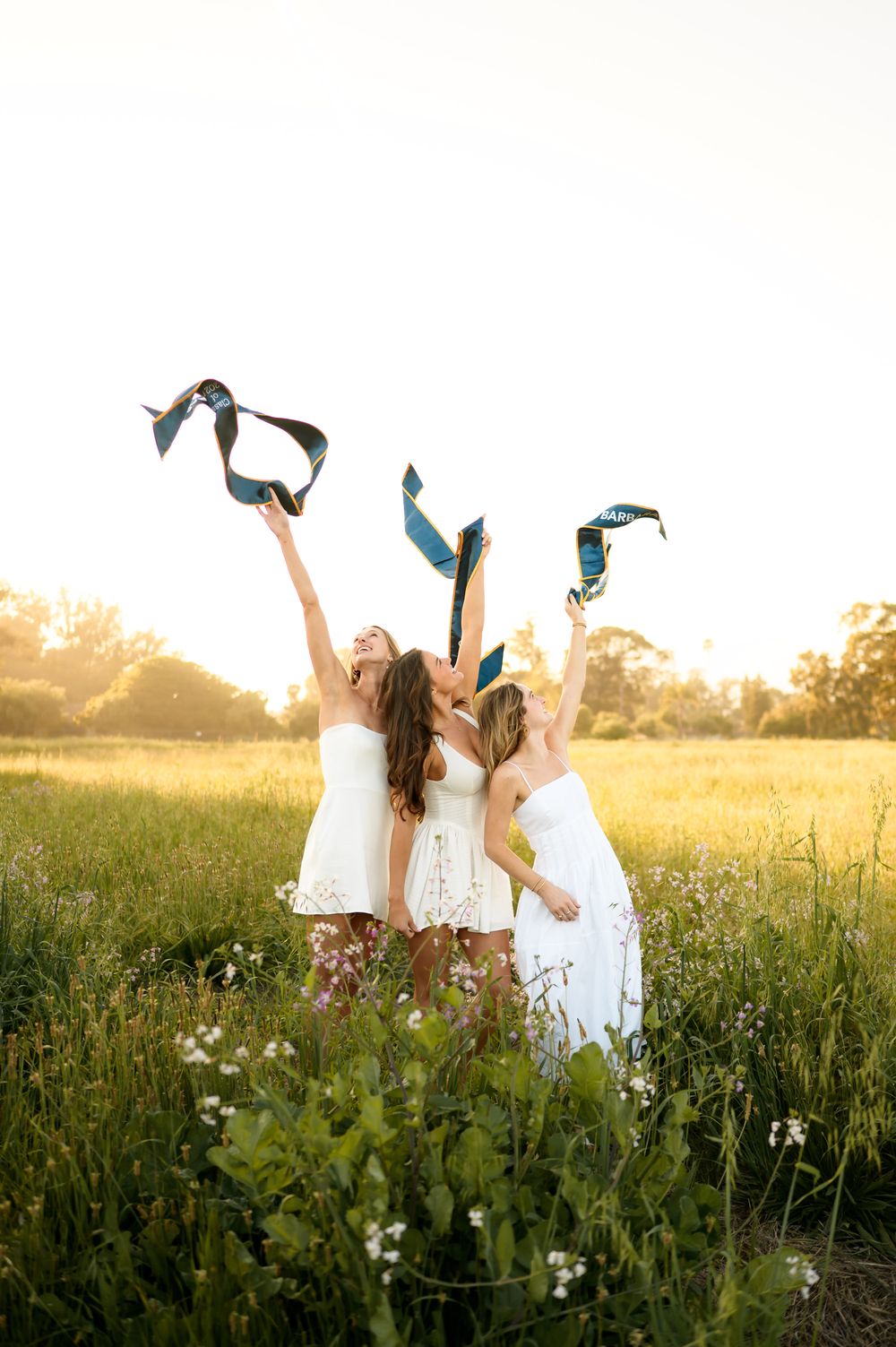 UCSB group grad portraits at Devereux Beach flower field during sunset of sorority seniors in white dresses holding up grad stoles