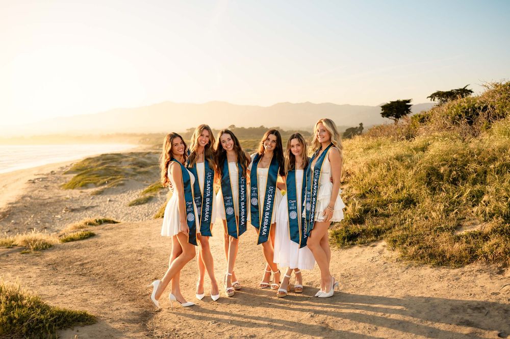 UCSB group grad portraits at Devereux Beach during sunset of sorority seniors in white dresses wearing grad stoles surrounded by sunlight