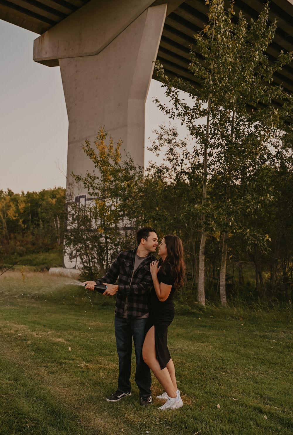 A couple shares a romantic moment under a bridge at sunset in a grassy outdoor setting.
