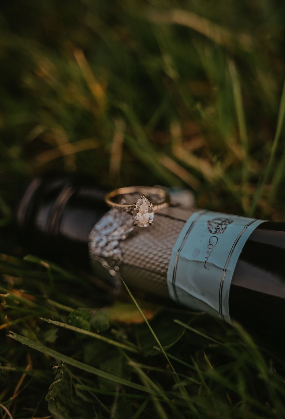 Diamond engagement ring rests on a wine bottle nestled in green grass during golden hour.