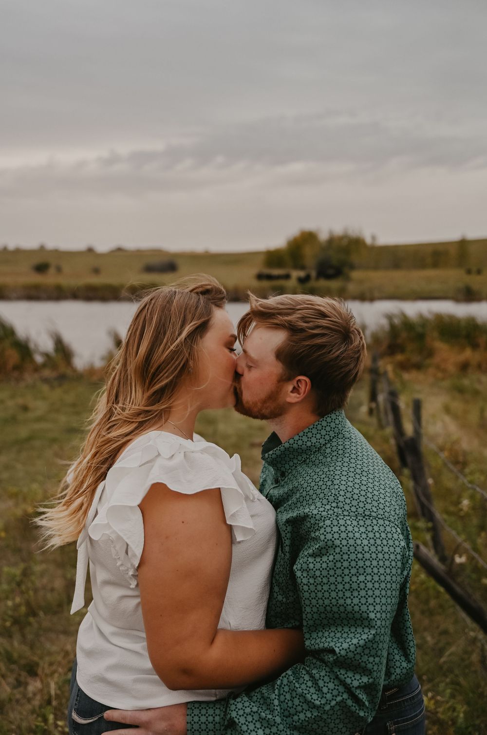 A romantic couple sharing a tender kiss by a lake on an overcast autumn day.
