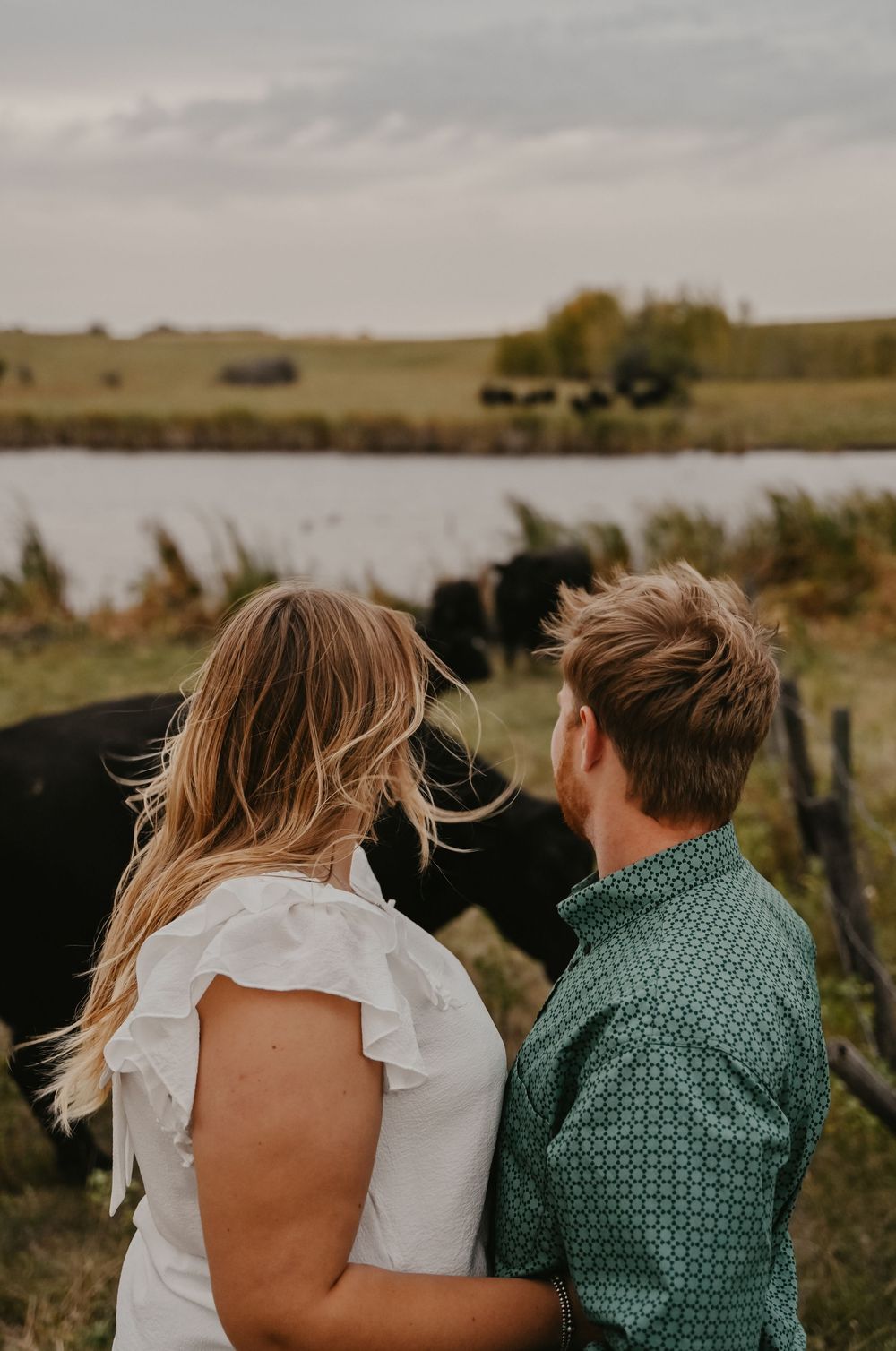 A couple in casual attire stands together against a scenic rural landscape at sunset.