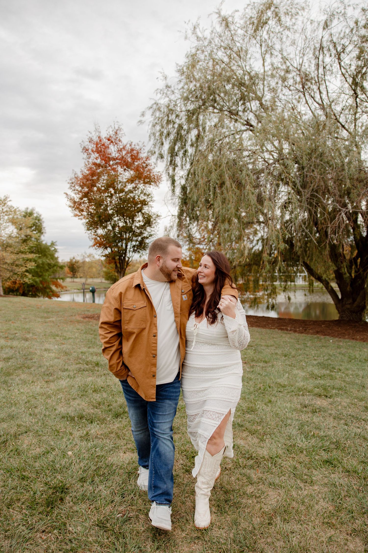 A couple walks together on grass near a lake with autumn trees and weeping willows.
