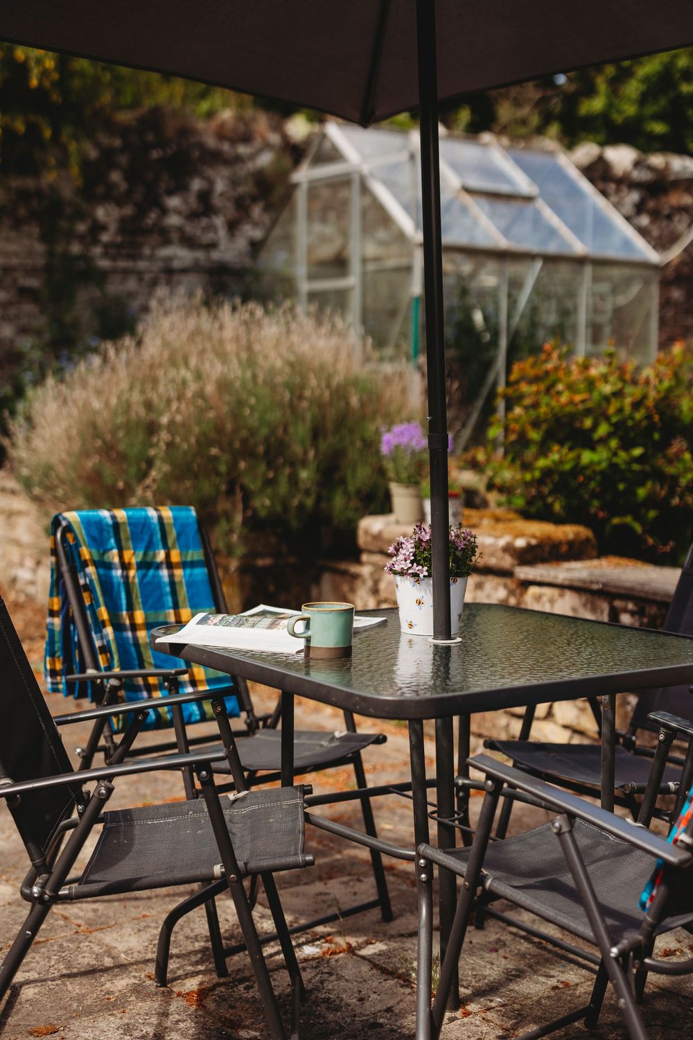 Outdoor patio setting with plaid chairs and umbrella table against a garden backdrop with greenhouse.