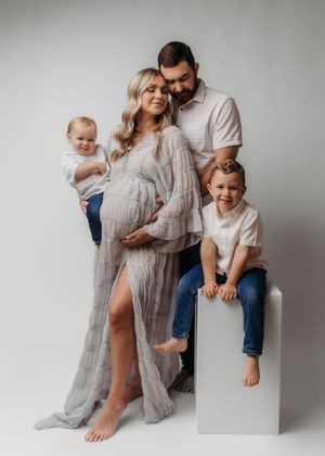 Family maternity photo with pregnant woman in gray dress, husband, toddler son, and baby in a bright studio setting.
