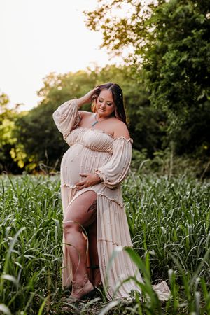 Pregnant woman in flowing off-shoulder beige gown standing in lush green field, cradling baby bump during golden hour maternity shoot.
