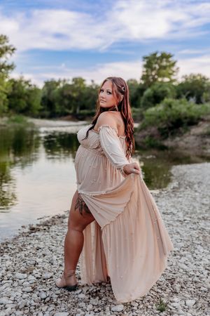 A woman in a flowing beige off-shoulder dress poses gracefully by a rocky riverbank surrounded by lush green trees.