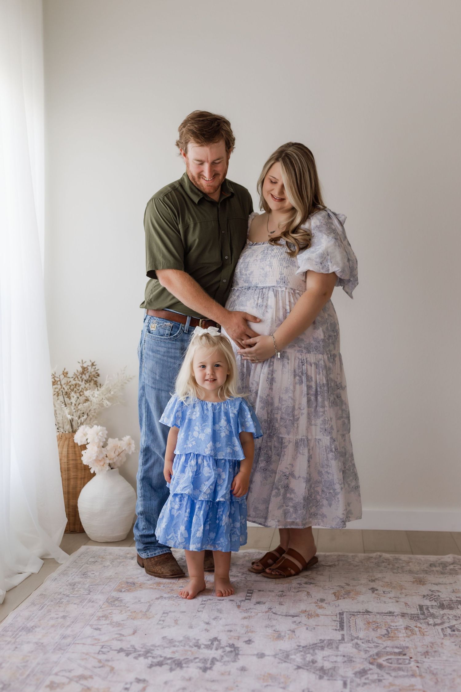 Family maternity photo with pregnant woman, man, and toddler girl in blue dress posing together indoors.