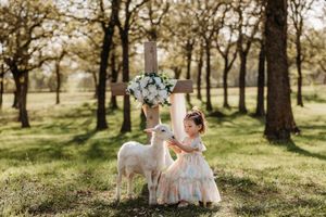 Young girl in floral dress feeding two white lambs near a wooden cross decorated with flowers outdoors.
