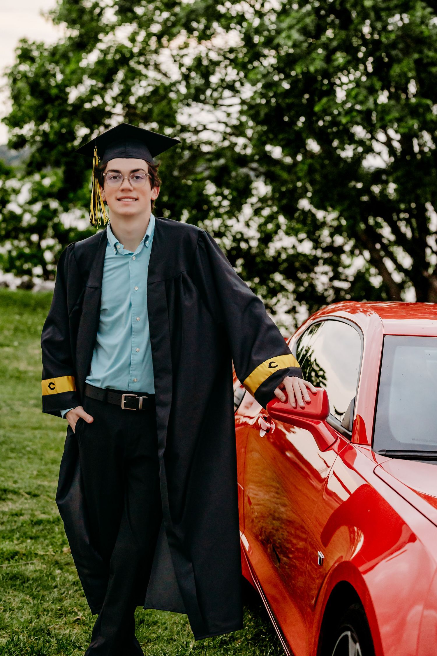 Young man in black graduation cap and gown leaning on a red car outdoors, smiling proudly on graduation day.