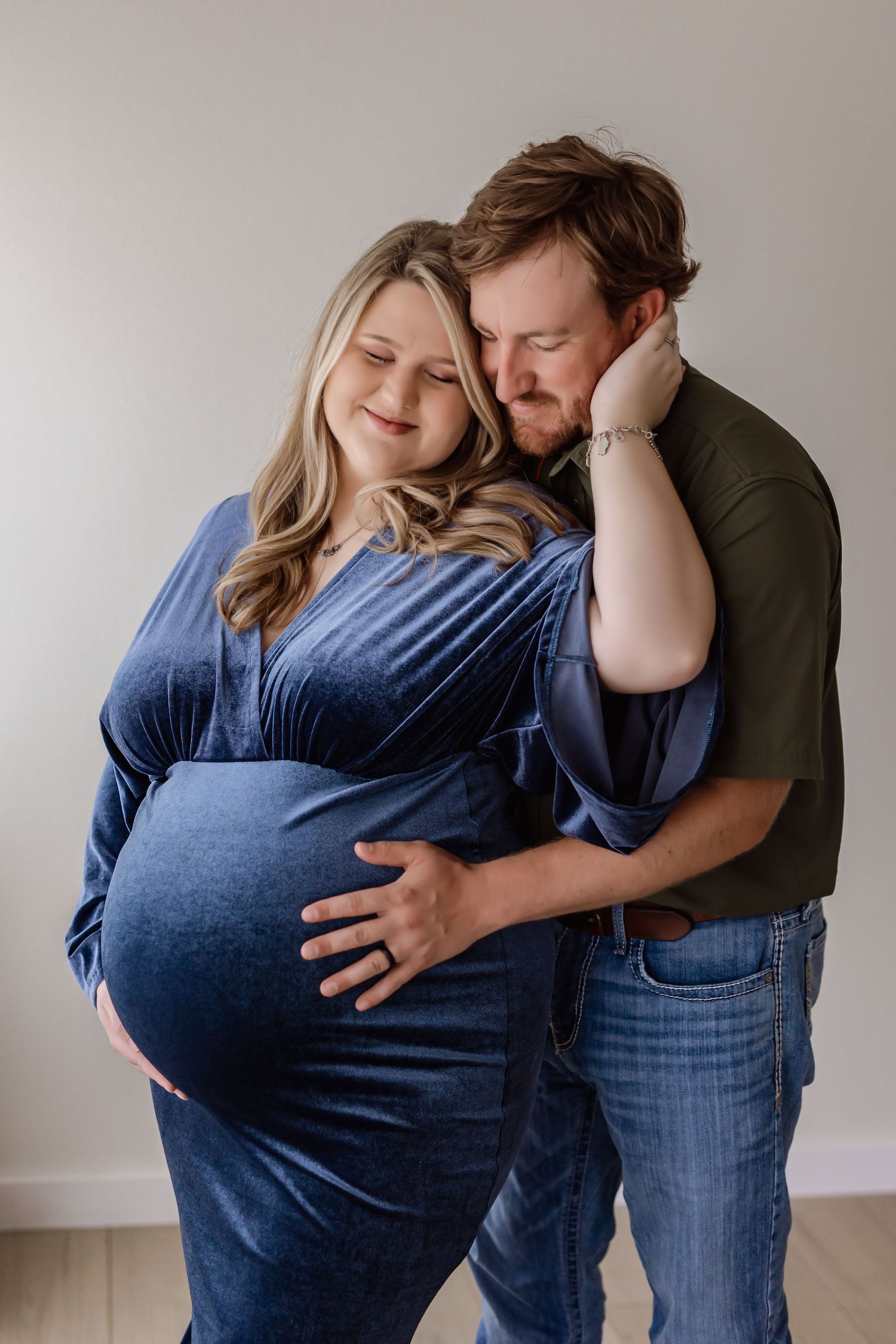 Couple sharing tender maternity photo moment, pregnant woman in blue velvet dress embraced by man in olive shirt.