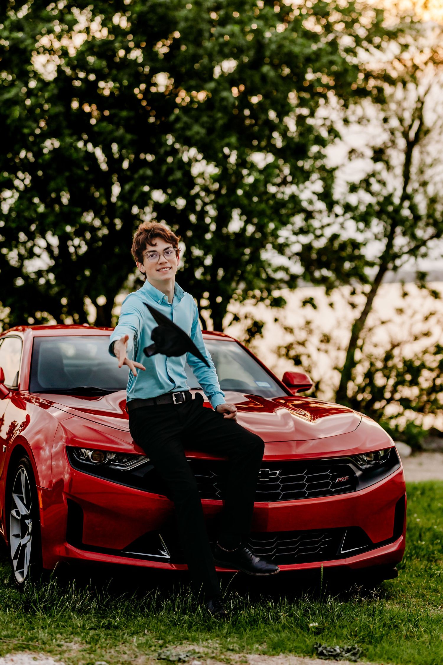 Young man in glasses poses leaning on a red Chevrolet Camaro SS outdoors at sunset.