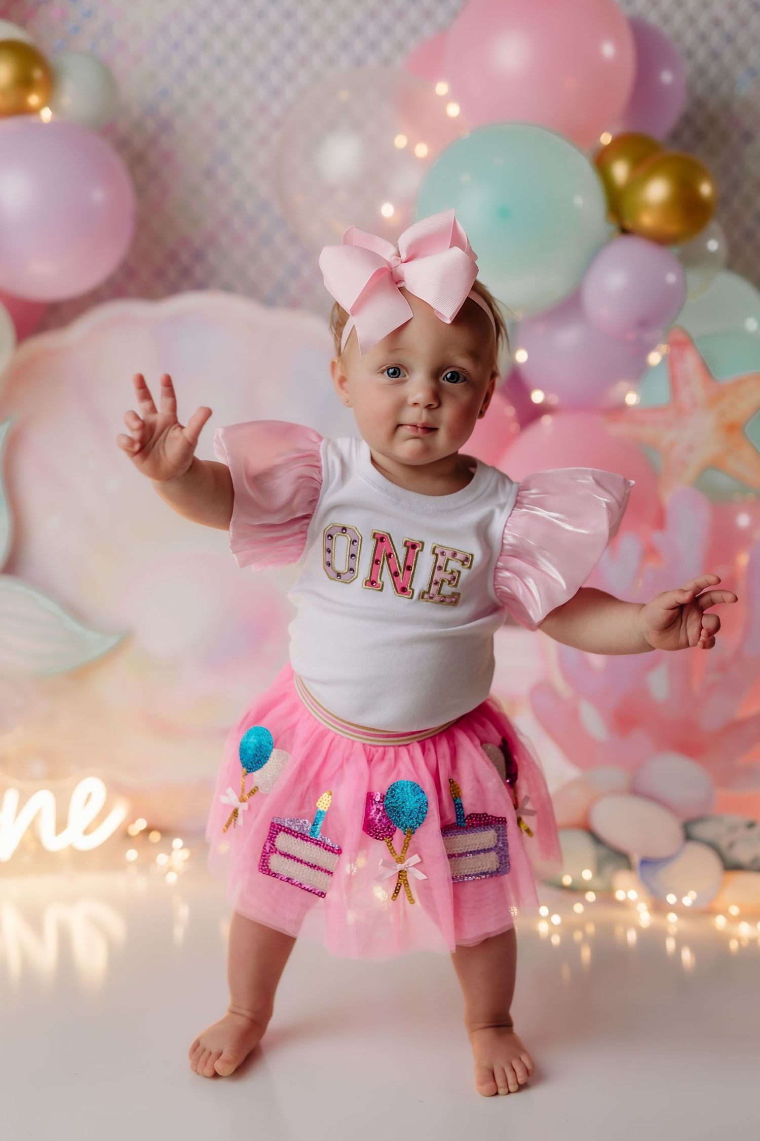 Baby girl in pink tutu and ONE shirt celebrates first birthday with colorful balloon backdrop and fairy lights.