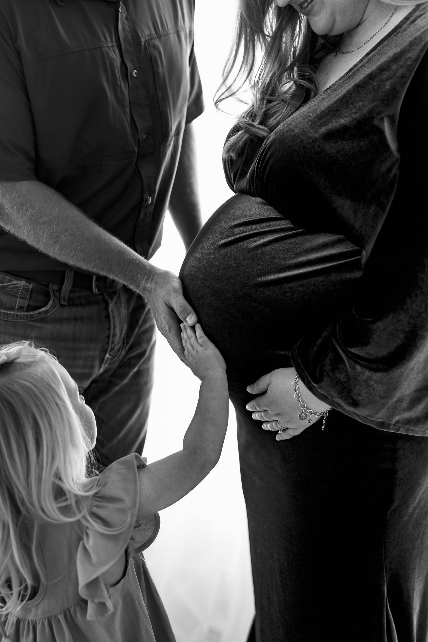 Black and white maternity photo of pregnant woman, man, and young girl touching baby bump together.