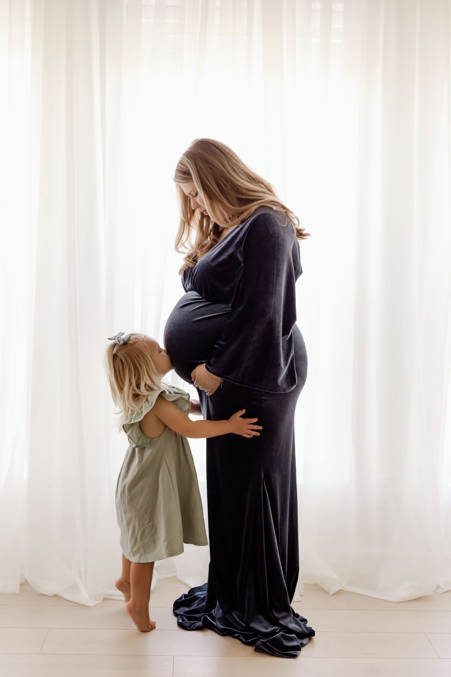 Pregnant woman in black dress with toddler girl touching her baby bump during maternity photo session.