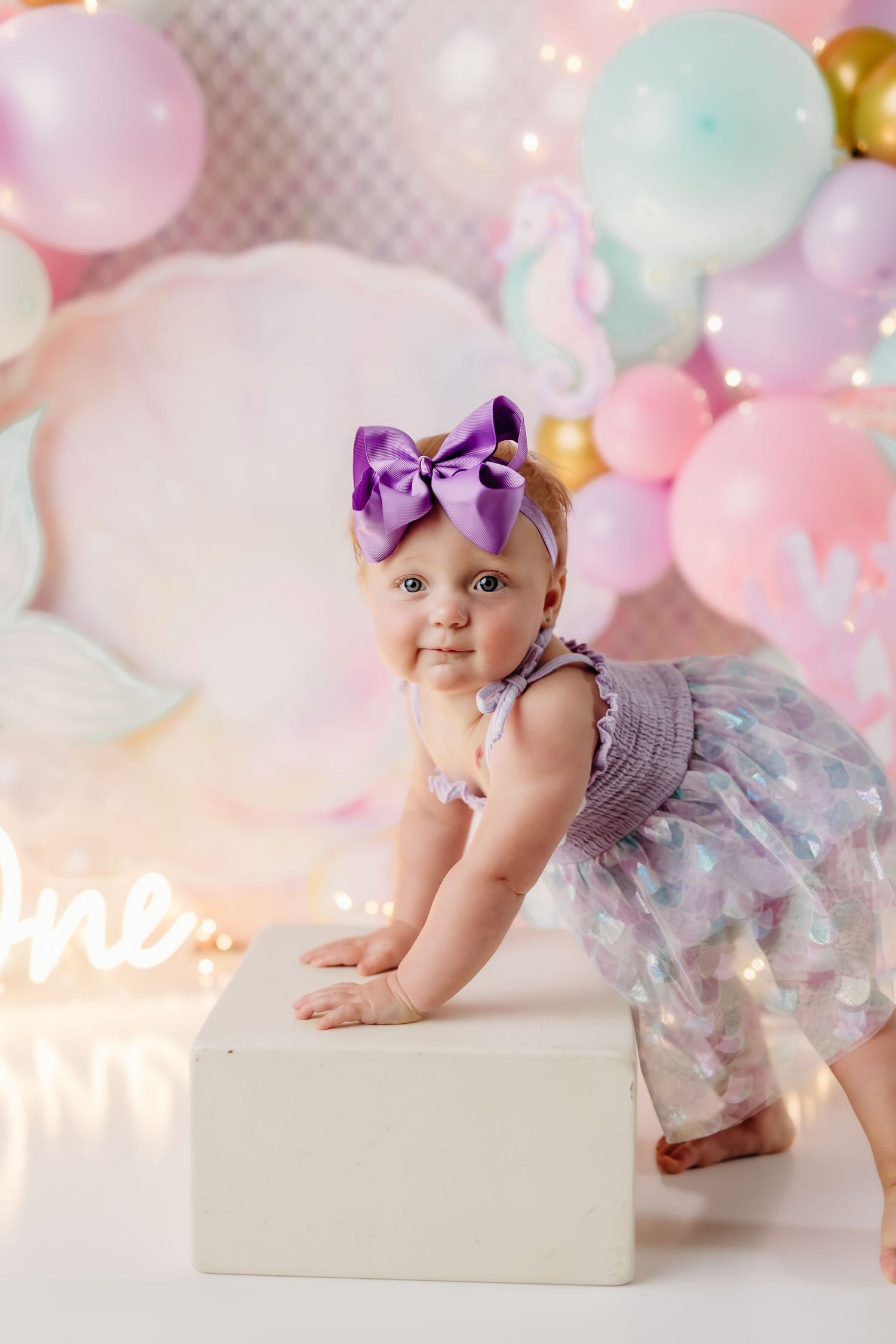 Baby girl in purple dress and bow headband poses by white box at unicorn-themed first birthday party with pastel balloons.