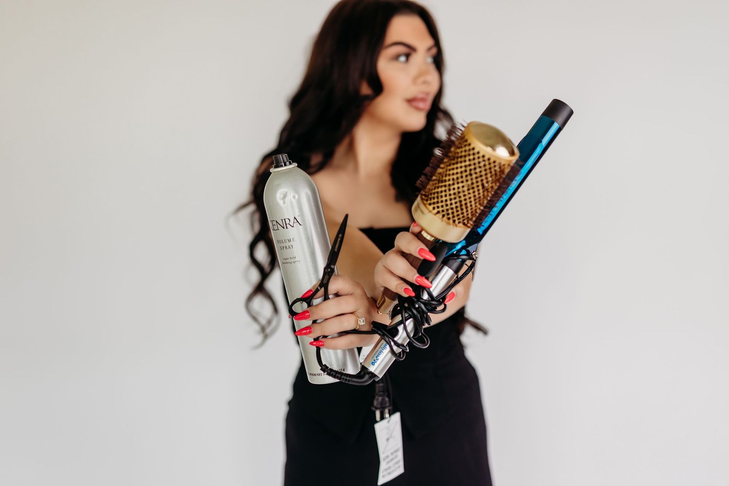 Woman in black outfit holding hair styling tools including a round brush, flat iron, and INOA hair product.