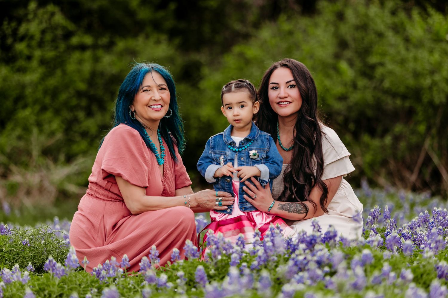 Three women sit in a field of purple bluebonnets, smiling together in a lush green outdoor setting.