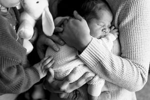 Black and white photo of newborn baby cradled in parents arms with a stuffed bunny toy nearby.