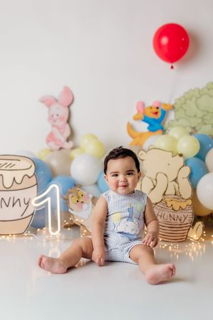 Baby boy sits smiling at his first birthday party with Winnie the Pooh decorations, balloons, and a glowing number one.