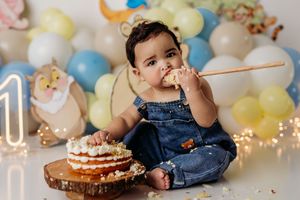Baby in denim overalls eating cake at a first birthday smash cake photoshoot with balloon backdrop.
