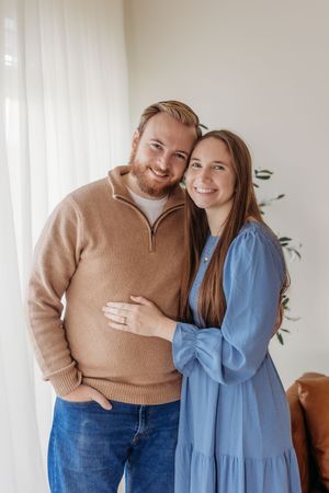 Happy couple smiling together indoors, man in beige sweater and woman in blue dress embracing near bright window.