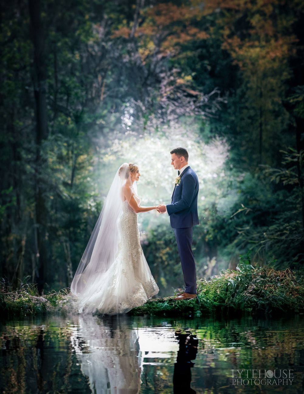 A romantic wedding photo captured in a misty forest setting beside a reflective pond with ethereal lighting.