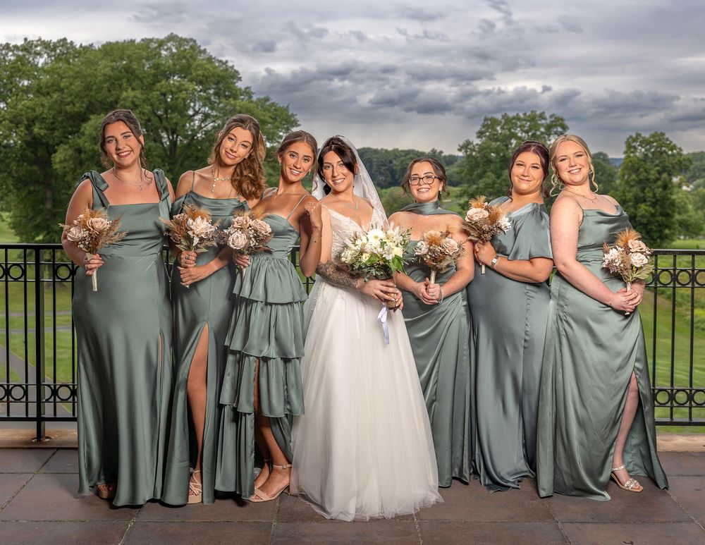 Bride in white gown poses with six bridesmaids in sage green dresses holding bouquets on an outdoor terrace.