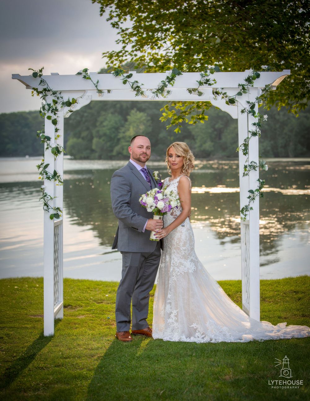 Bride and groom pose under white floral arbor by lakeside at sunset wedding ceremony.