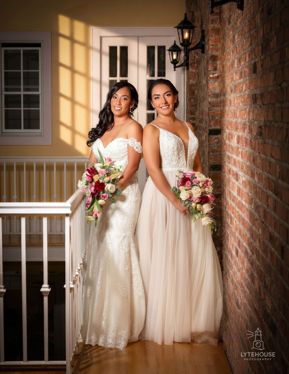 Two brides in white wedding gowns holding floral bouquets pose elegantly on a staircase with brick walls.