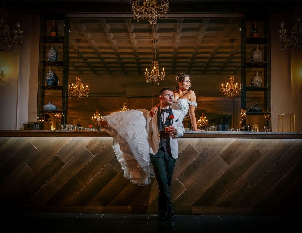 Groom lifting bride in elegant wedding venue with chandelier and wooden bar backdrop.