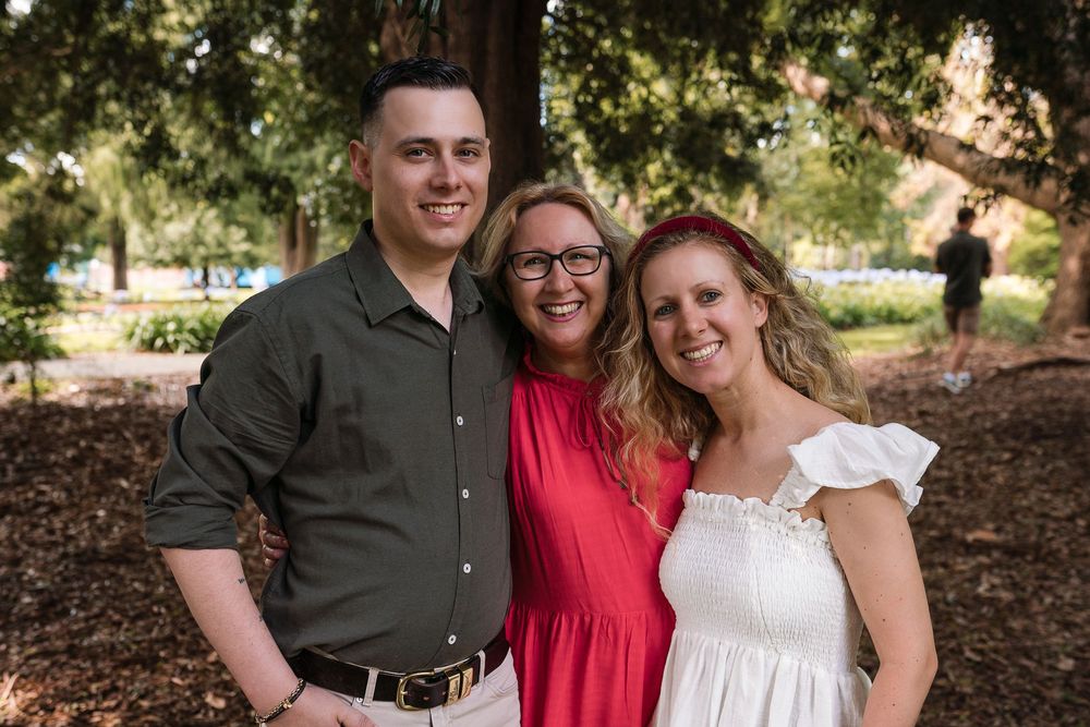 A group of three people standing together outdoors. A man on the left is wearing a dark shirt, a woman in the middle is wearing a red dress,