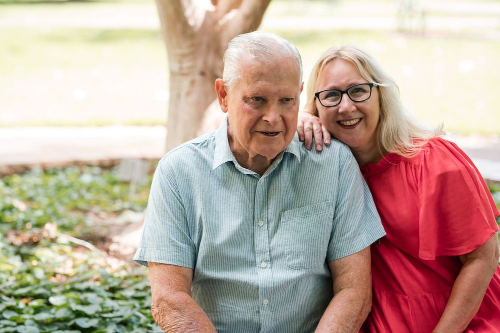 An elderly man in a light blue shirt is sitting outdoors, with a woman in a red dress standing beside him, smiling. They are in a garden set