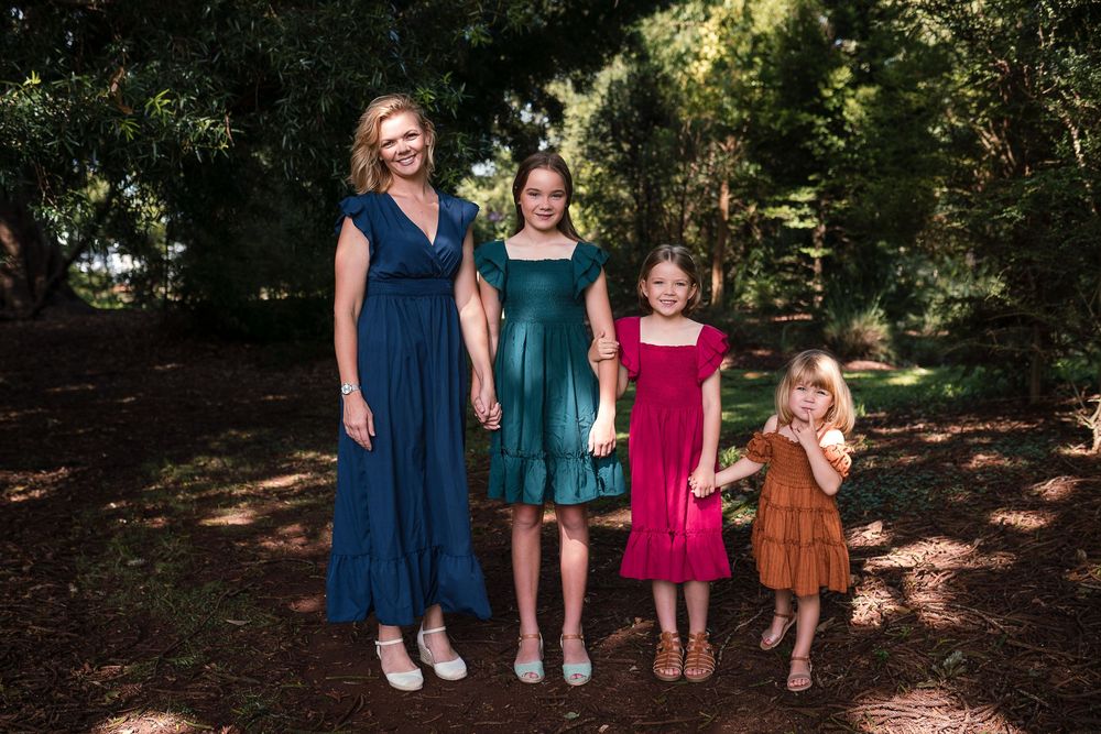 A woman and three young girls are standing together outdoors in a wooded area. The woman is wearing a blue dress, and the girls are wearing