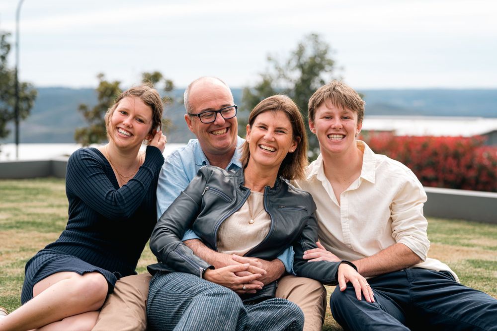 A family four sitting & grass, smiling. The group includes two adults and two young adults. They are outdoors with a scenic view  background