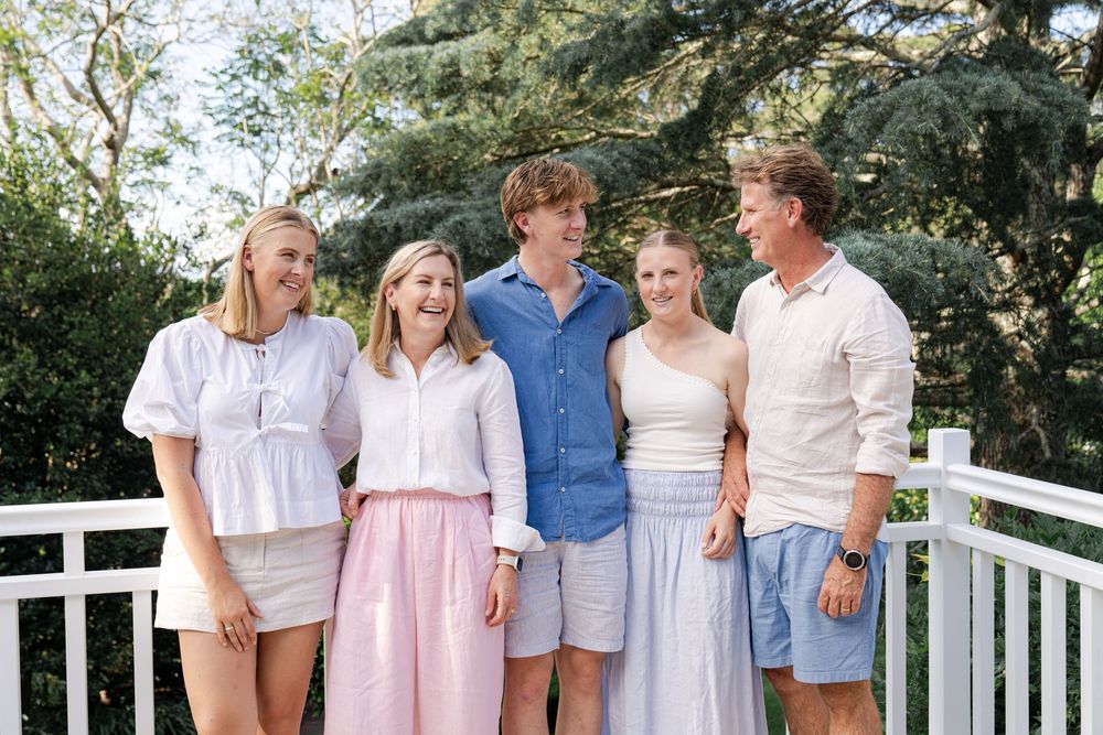 A group of five people standing on a porch, smiling at each other. They are dressed in light, casual summer clothing. There are in trees  us