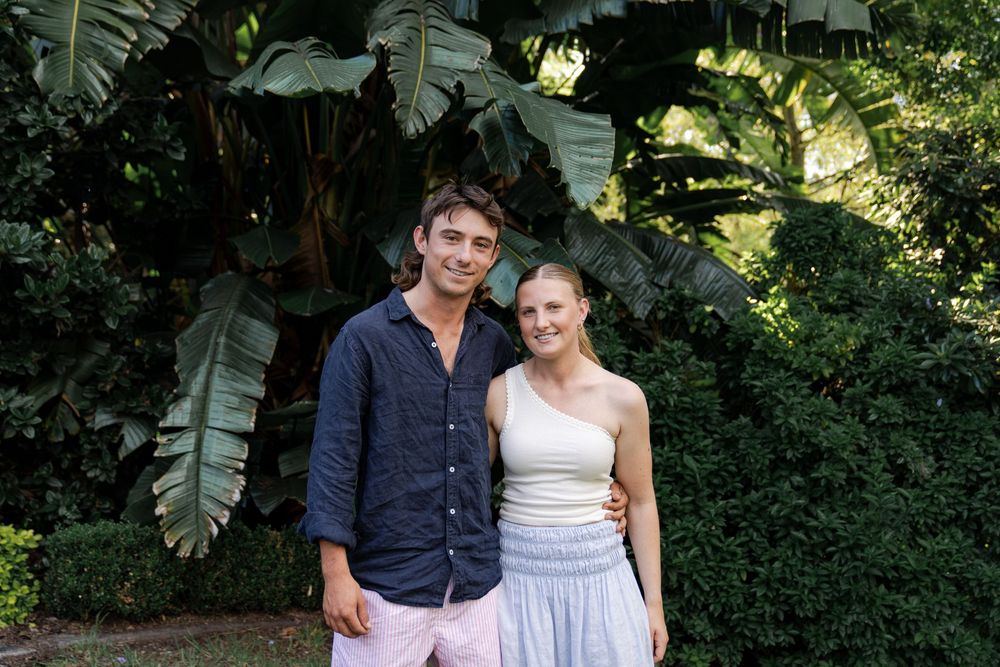 A man and a woman are standing together outdoors  front  large green plants. The man is wearing a dark shirt and light shorts, and the skirt