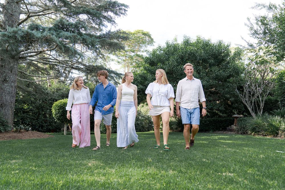A group of five people walking on grass in a garden. They are casually dressed appear to be enjoying  sunny day. Trees and bushes background