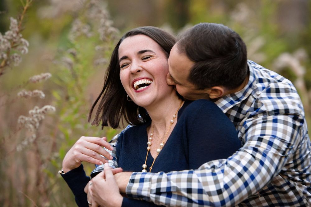 Romantic couple embraces and laughs while taking outdoor engagement photos in a natural setting.