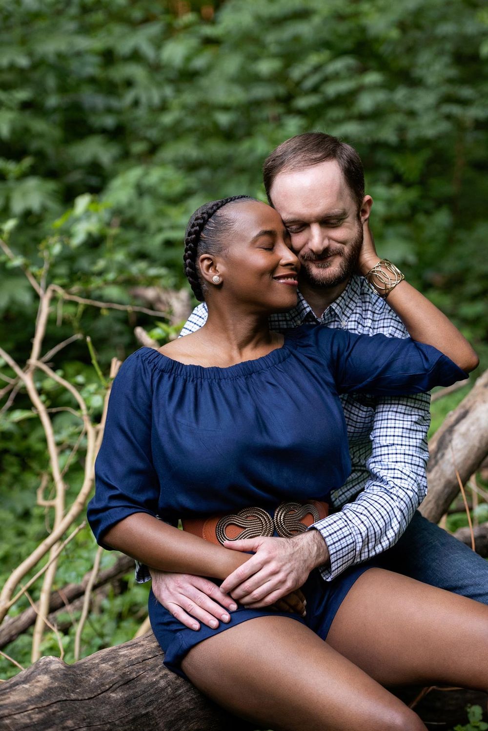 Interracial couple sharing intimate moment while sitting on fallen tree in forest setting.