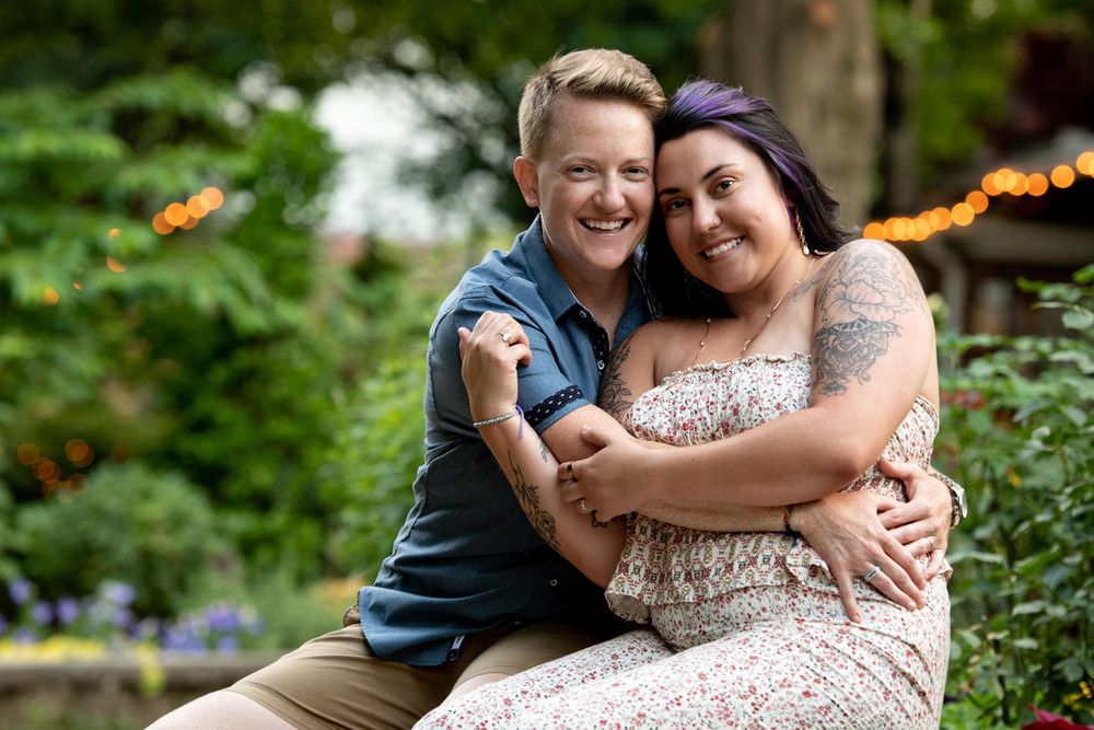 Two people share a warm embrace while sitting outdoors surrounded by greenery and flowers in a garden setting.