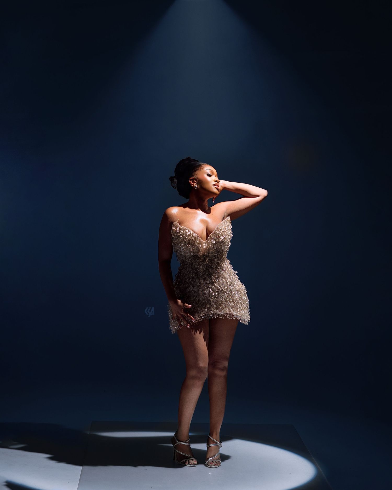 Woman in sequined mini dress poses dramatically under spotlight on dark blue stage background.